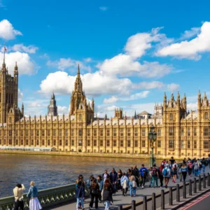 Crowds walking along the Thames with the Palace of Westminster under a blue sky